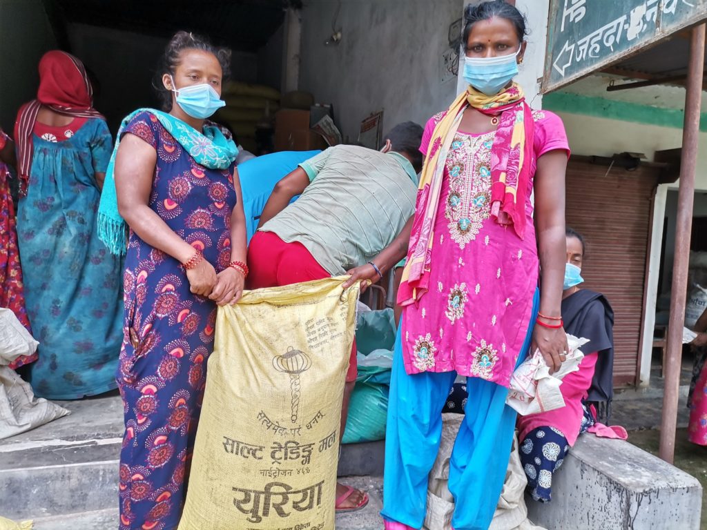 Two Nepalese women in colorful garments and temporary masks hold a shared bag, standing in front of an outdoor aid distribution center.