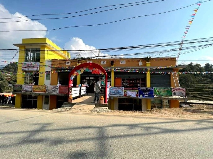 In this photo, we see a very colorful one-story building across the street. It is yellow and orange, and adorned with Nepalese Prayer Flags, as well as an arch of balloons around the entrance, celebrating the Center's recent opening. Power lines cross the sky in the foreground, and a forest of trees colors the distant in the background behind the Center. 